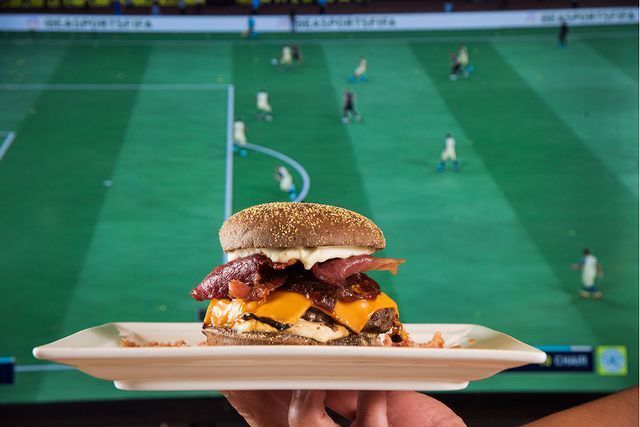 pessoa segurando um prato com hamburguer em frente uma tv com jogo de futebol