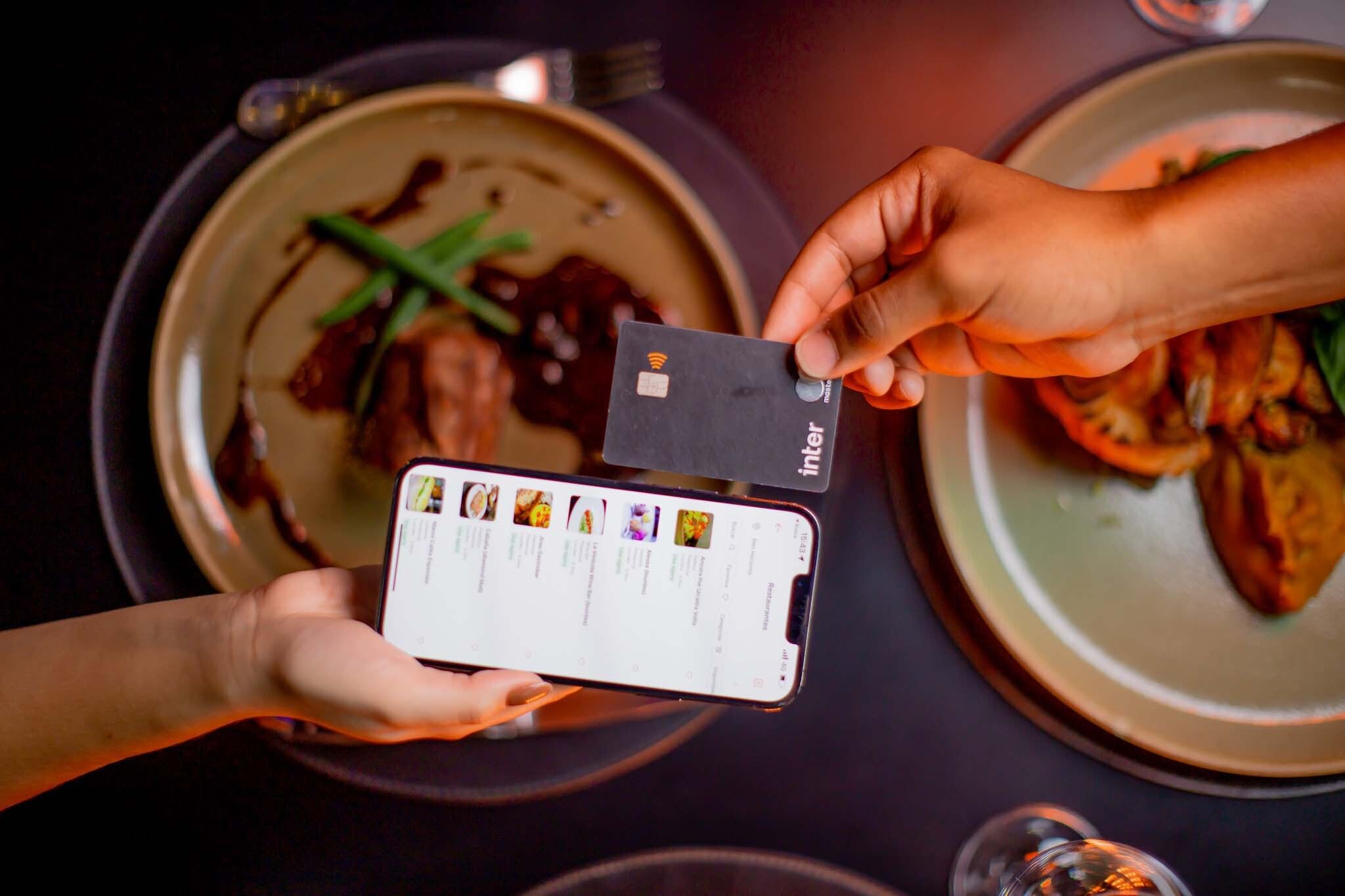 Mão segurando um celular com o app Duo Gourmet na tela e outra mão segurando um cartão Black com 2 pratos de comida ao fundo