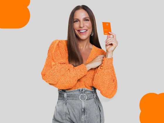 Foto da cantora Ivete Sangalo sorrindo, vestindo calça jeans e blusa laranja, segurando um cartão laranja do Inter