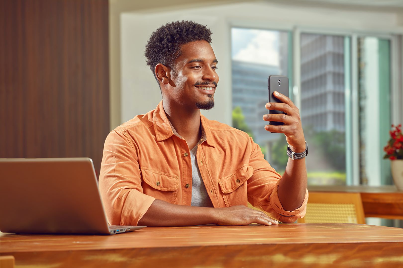 Homem jovem sorrindo, vestindo uma blusa laranja, sentado de frente a uma mesa com computador e celular em mãos,  como se estivesse navegando pelo Super App do Inter.