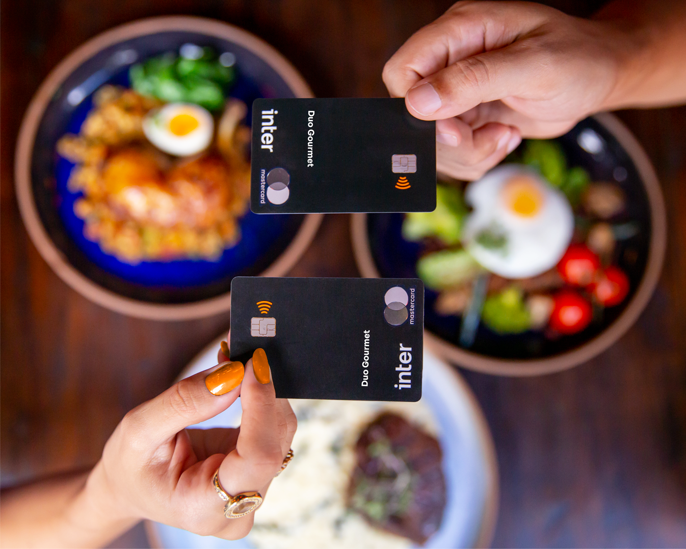 Imagens duas mãos segurando seus cartões Inter Black, com pratos de comida ao fundo, simbolizando o pagamento com Duo Gourmet
