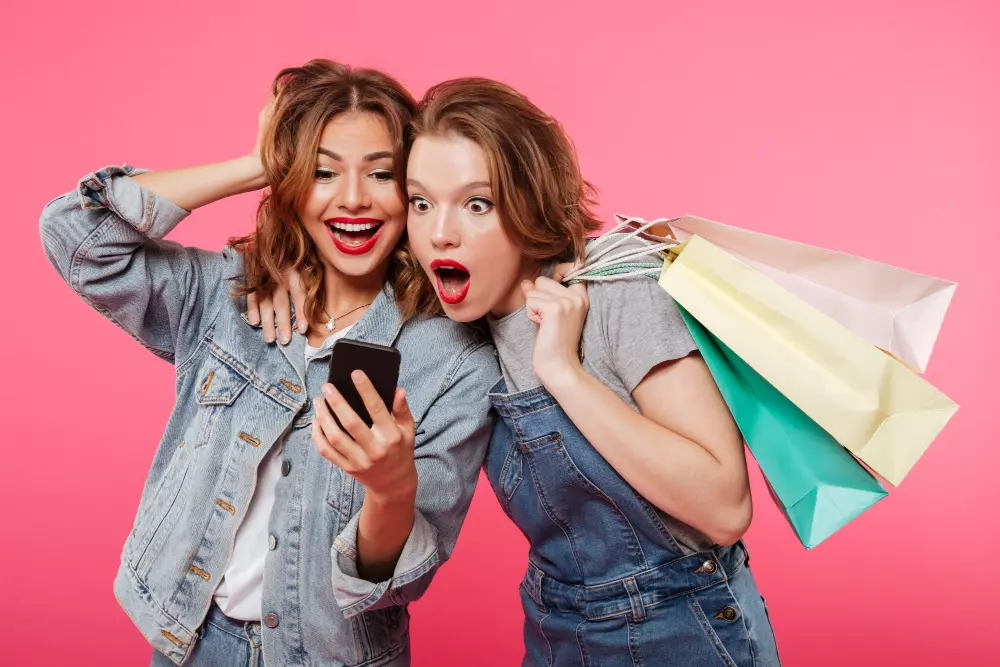 Duas mulheres com rosto surpreso olhando para a tela do celular e uma delas segurando sacolas de compras. 
